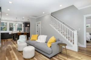 Cozy living room area with a grey sofa, yellow accent pillows, polished hardwood floors, and a white open staircase.