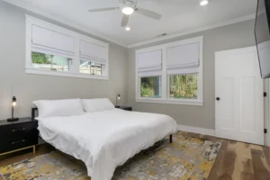 Bright bedroom interior with light grey walls, a white bed, dual windows with privacy blinds, and a patterned area rug.