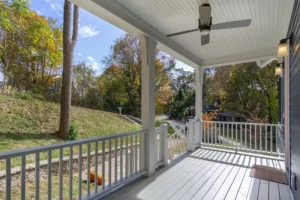 Covered front porch view looking out from a custom home, featuring a painted light grey wood deck, clean white railings and columns, a white beadboard ceiling with a modern ceiling fan, and dark blue exterior siding.