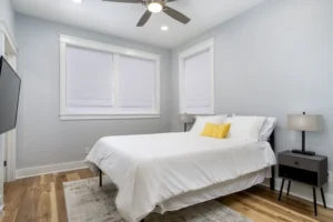 Cozy bedroom interior with grey walls, a white bed with a yellow pillow, and windows with white blinds.