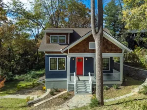 Front exterior shot of a blue craftsman house framed by trees, highlighting a white porch and red door.