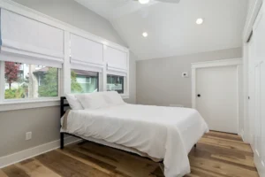 Bright master bedroom interior with a white bed setup, vaulted ceiling, and large windows with privacy blinds.