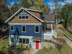 Side exterior view of a modern blue craftsman house highlighting a bright red door and yellow outdoor chairs.
