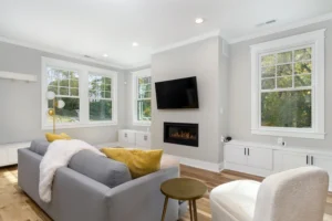 Cozy living room interior featuring a light grey sofa, a modern built-in fireplace, a wall-mounted TV, and large windows.