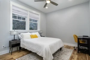 Sunlit master bedroom featuring clean grey walls, a white bed setup with a yellow accent pillow, and large windows with blinds.