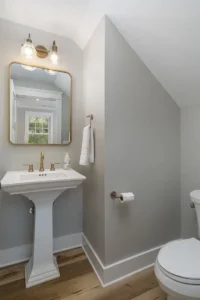 Minimalist powder room featuring a classic white pedestal sink, a gold rectangular mirror, and light grey walls.