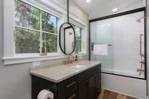 Modern bathroom vanity area with a dark cabinet, premium stone countertop, a round black-rimmed mirror, and a glass shower.