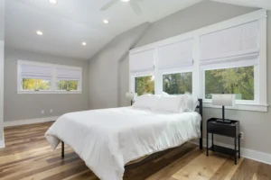 Bright master bedroom interior featuring a white bed setup, high ceilings, and large multi-pane windows.